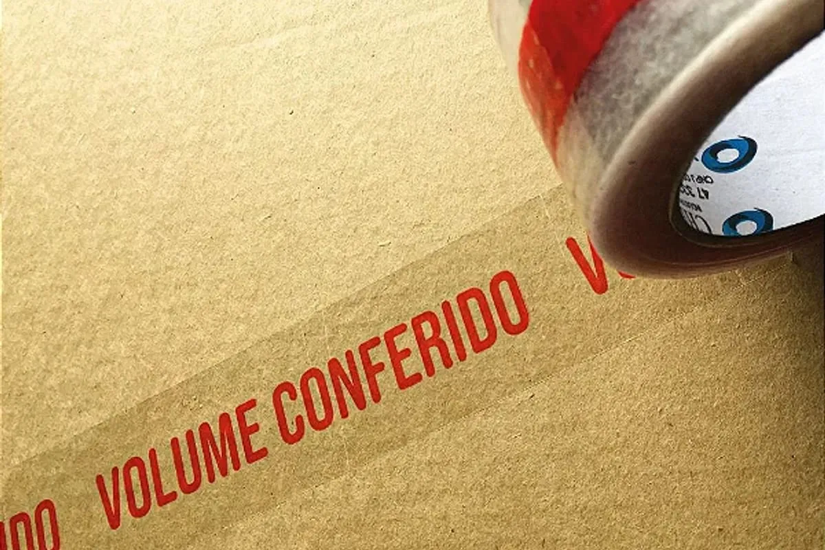Fita adesiva com a mensagem VOLUME CONFERIDO impressa em vermelho sobre caixa de papelão kraft